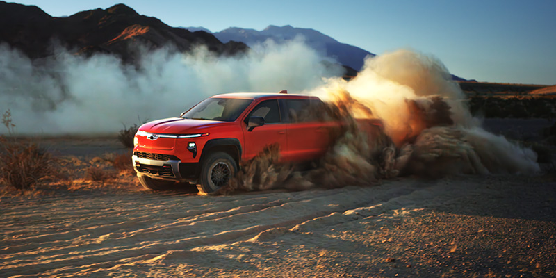 Red electric pickup truck kicking up dust while driving off-road through a desert landscape with mountains in the background.