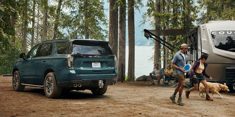 Full-size SUV parked at a lakeside campsite with a family, dog, and camper enjoying an outdoor adventure among tall trees.