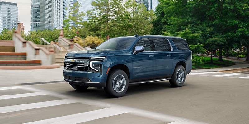 Blue Chevrolet Suburban driving through city streets with trees and buildings